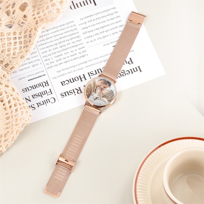 Peut inclure: Une montre &agrave; bracelet en maille dor&eacute;e rose avec un cadran rond pr&eacute;sentant une photo d'un couple. La montre est sur une surface blanche avec un morceau de tissu en dentelle en arri&egrave;re-plan.