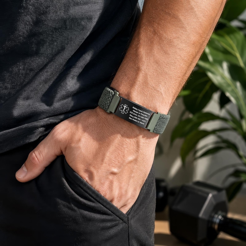 May include: An olive green medical alert bracelet featuring a woven fabric band and a black rectangular metal plate. The plate displays a white Star of Life symbol and engraved text detailing medical information and emergency contact numbers.