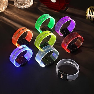 Op de afbeelding: Een verzameling verlichte polsbandjes in verschillende kleuren, waaronder oranje, groen, paars en rood. Elke band heeft gepersonaliseerde tekst zoals "Love you forever" en namen met data. De bandjes hebben een zwarte rechthoekige basis.
