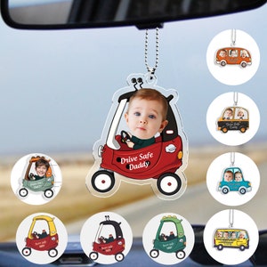 Pode incluir: Um carro de brinquedo vermelho, branco e preto com o rosto de uma criança no banco do motorista. O carro tem as palavras "Drive Safe Daddy" no lado. O carro está pendurado em um espelho retrovisor com uma corrente prateada.
