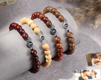 Puede incluir: Se muestran cuatro pulseras de cuentas de madera en tonos marrones y beige. Cada pulsera presenta cuentas redondas de madera y una cuenta de acento de metal oscuro. Las pulseras están dispuestas sobre una superficie gris curvada, con dos fotos pequeñas en el fondo.