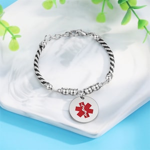 May include: A silver bracelet with a black and white braided cord and a silver charm with a red medical symbol.