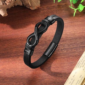 May include: Black leather bracelet with a matte black infinity symbol clasp. The bracelet has the names "Daine" and "Tania" engraved on it, with "Always and forever" inscribed on the infinity symbol. The bracelet has a magnetic clasp.