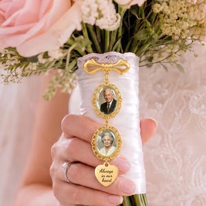 May include: A gold-toned metal wedding bouquet charm featuring a bow-shaped top, two oval frames containing portraits of an older man and an older woman, and a heart-shaped pendant engraved with the text Always in our heart.