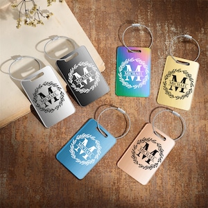 Op de afbeelding: Zes gepersonaliseerde bagagelabeltjes met een kransontwerp en de naam "MICHAEL" gegraveerd op elk labeltje. De labeltjes zijn in verschillende kleuren verkrijgbaar: zilver, zwart, regenboog, blauw en rosé goud.
