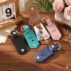 May include: A collection of personalized leather keychains in various colors, including black, teal, pink, and navy blue. Some keychains feature names like "Carlos" and "Daddy," while others have text like "My first hero, Best dad ever."