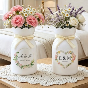 May include: Two white ceramic vases with scalloped rims, each tied with a cream ribbon. One vase holds pink carnations and daisies, the other lavender, daisies, and white roses. Each vase has a floral design and initials with a date.