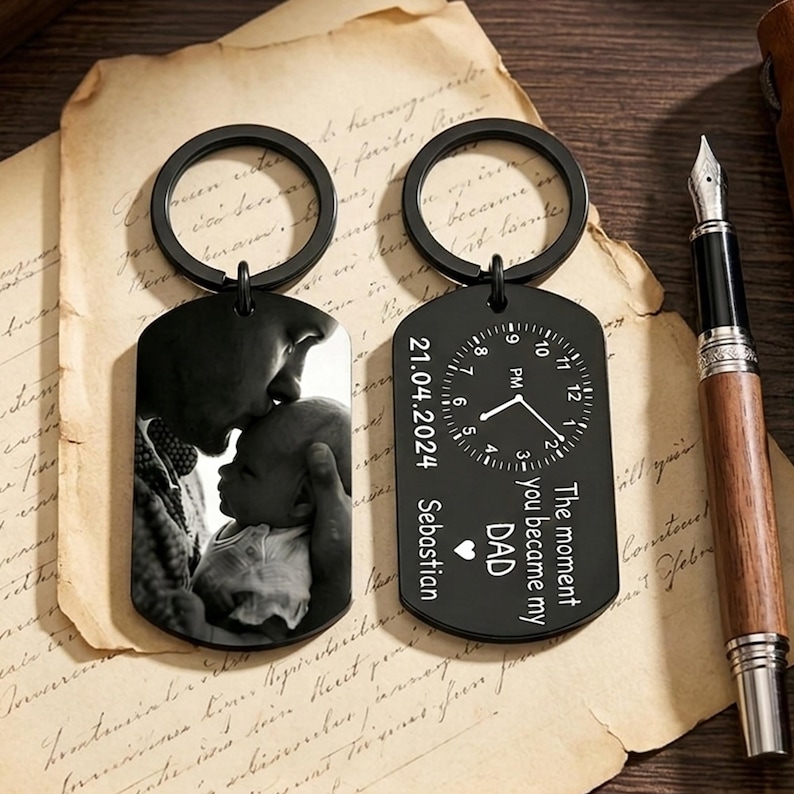 May include: Two black keychains. One features a black and white photo of a person kissing a baby. The other has a clock design with the text "The moment you became my DAD" and the date 21.04.2024, with the name Sebastian.