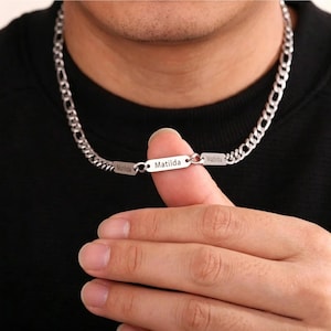 May include: A silver-colored chain necklace with a rectangular pendant that reads "Matilda". The necklace has a curb chain design and is worn around the neck. The name "Matilda" is also engraved on two smaller rectangular pendants.