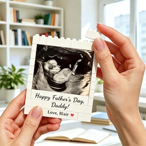 Peut inclure: Cadre photo blanc en forme de bloc avec une image d'échographie. Le cadre porte l'inscription "Happy Father's Day, Daddy! Love, Blair" avec un symbole de cœur. Le cadre est tenu par une personne.