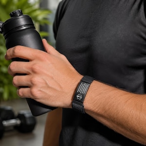 May include: A black medical alert bracelet featuring a textured silicone band and a rectangular metal plate engraved with a medical star of life symbol and personal identification text, worn on the wrist while holding a black water bottle.