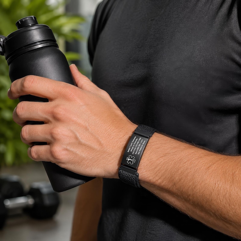 May include: A black medical alert bracelet featuring a textured silicone band and a rectangular metal plate engraved with a medical star of life symbol and personal identification text, worn on the wrist while holding a black water bottle.