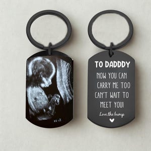 May include: Black keychain with a black ring. One side features a grayscale ultrasound image of a foetus. The other side has the text "TO DADDY NOW YOU CAN CARRY ME TOO CAN'T WAIT TO MEET YOU! Love, the bump."