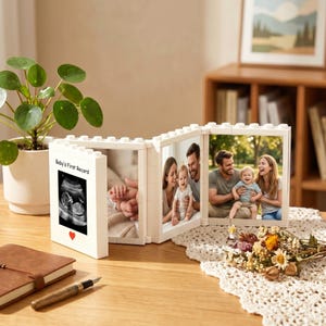 Könnte beinhalten: Ein weißer, modularer, klappbarer Bilderrahmen im Baustein-Design mit vier Bildern: ein Baby-Ultraschallbild mit der Aufschrift 'Baby's First Record' und einem roten Herz, Babyhände, ein Familienporträt und ein Paar mit einem Kleinkind.