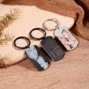 May include: Three personalized keychains with photos and text. One keychain has a black ring and a black tag with a photo of a man and a baby. Another has a black ring and a black tag with a clock design and text. The third has a silver ring and a photo.