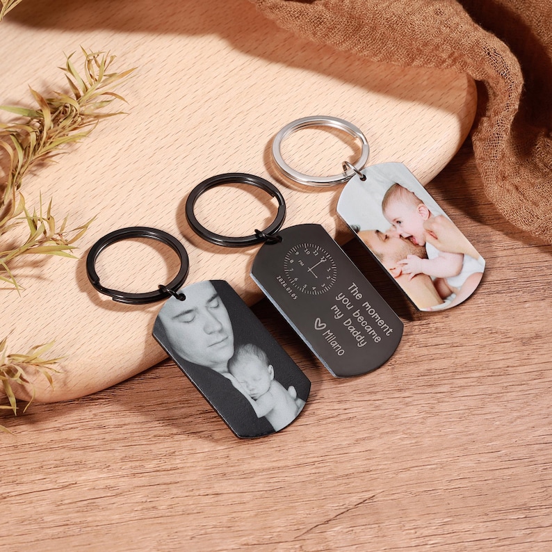 May include: Three personalized keychains with photos and text. One keychain has a black ring and a black tag with a photo of a man and a baby. Another has a black ring and a black tag with a clock design and text. The third has a silver ring and a photo.