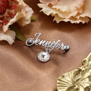 May include: Silver-toned name pin with the word "Jennifer" in cursive script. A clear crystal charm dangles from the pin.