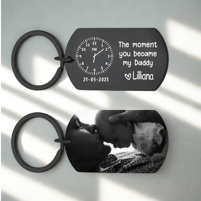 May include: Black metal keychain with two rectangular pendants. One pendant features a clock design with the text "The moment you became my Daddy Lilliana" and the date "21-05-2023". The other pendant displays a black and white photo.