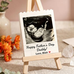 Peut inclure: Un cadre photo blanc en forme de bloc avec une image d'échographie, présenté sur un petit chevalet en bois. Le cadre porte l'inscription "Happy Father's Day, Daddy! Love, Blair" en noir avec un cœur rouge.