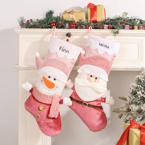 Könnte beinhalten: Zwei rosa Weihnachtsstrümpfe mit personalisierten Namen, "Finn" und "Meyra". Ein Strumpf zeigt ein Schneemann-Design, während der andere ein Weihnachtsmann-Design hat.