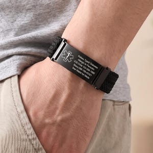 Puede incluir: Una pulsera de alerta médica negra con una banda de nailon tejida y una placa metálica rectangular. La placa muestra un símbolo de la Estrella de la Vida en blanco y el texto grabado: Kelsey: Type2 Diabetes, Allergic to Penicillin, ICE:Chris 123-456-7890, Stacy.099-765-43321, See Wallet for Meds.