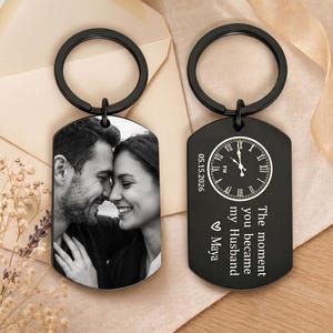 May include: Two black metal dog tag keychains with circular rings. One keychain features a black and white photograph of a couple. The other keychain displays a clock face, the date 05.15.2026, and the text: The moment you became my Husband ♡ Maya.