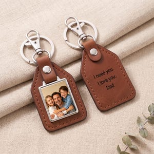 May include: Two brown leather keychains with silver metal hardware. One keychain features a square photo insert showing a man, a girl, and a boy smiling. The other keychain is engraved with the text: I need you. I love you. Dad.