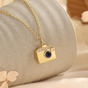 May include: A gold-colored camera pendant necklace. The camera pendant has a blue gemstone lens and is adorned with small, clear gemstones. The necklace chain is also gold-colored. The pendant is approximately 0.75 inches wide.