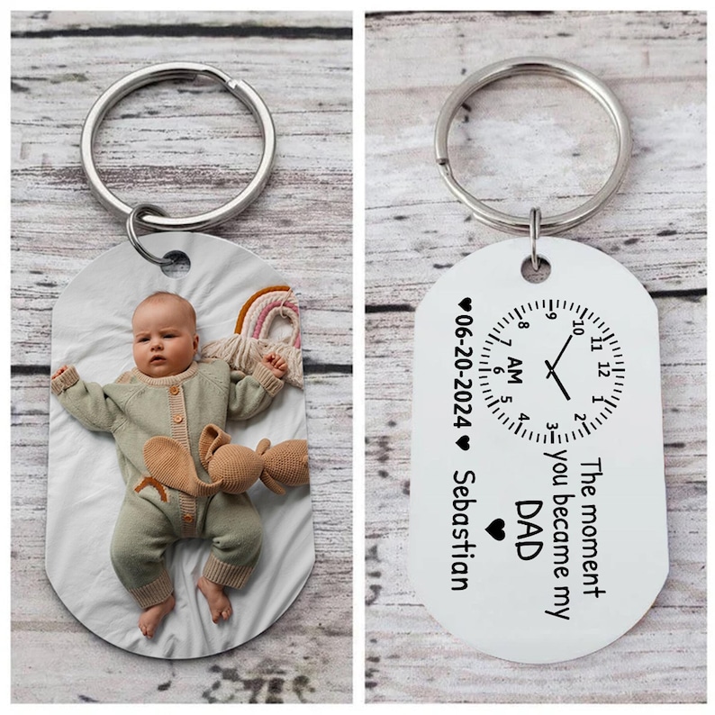 May include: A silver keychain with two tags. One tag features a baby in a green outfit, the other has a clock design with the text "The moment you became my DAD" and the date "06-20-2024".