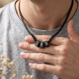 以下が含まれることがあります： 黒いキューブペンダントが3つ付いた黒いカーブチェーンネックレス。「12.22M」、「Mike」、「Lucy」と名前が刻まれています。ネックレスは首にかけられています。