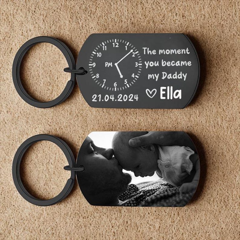 May include: Black keychain with a clock design and the text "The moment you became my Daddy" and the name "Ella". The second keychain has a black and white photo of a baby and a person.