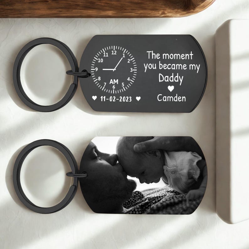 May include: Two black keychains. One keychain features a clock design with the text "The moment you became my Daddy Camden" and the date 11-02-2023. The other keychain displays a black and white photo of a baby and a person.
