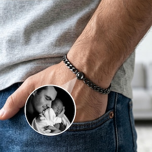 May include: A black chain bracelet with a circular clasp is shown on a wrist. A small, black and white photo of a man holding a baby is in the lower left corner. The bracelet is made of interlocking links.