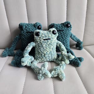 May include: Three crocheted frog plushies in shades of green. The frogs are sitting on a light gray couch.