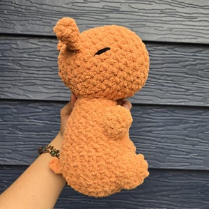 May include: A crocheted plush toy in the shape of a small animal. The toy is a light orange color and has a fluffy texture. The toy is being held by a hand.