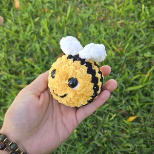May include: A handmade, plush bee toy with yellow and black stripes, white wings, and large black eyes. The bee is round and soft, held in a hand against a backdrop of green grass.