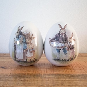 Può includere: Due decorazioni di uova di Pasqua in ceramica bianca. Ogni uovo presenta un'illustrazione dettagliata di conigli vestiti con abiti vintage, che trasportano cesti e ombrelli. Le uova sono su una superficie di legno.