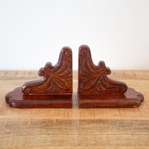 May include: Pair of dark brown wooden bookends with ornate, carved floral designs. Each bookend has a rectangular base and a vertical support with detailed leaf and scroll patterns. The bookends are displayed on a wooden surface.