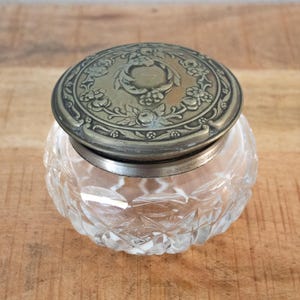 May include: A clear, cut-glass jar with a decorative, silver-toned lid. The lid features an embossed floral design. The jar sits on a wooden surface. The jar is likely used for storing small items.