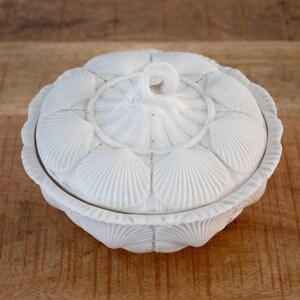 Puede incluir: Un cuenco redondo de cerámica blanca con tapa, que presenta una superficie texturizada con un patrón repetitivo de conchas marinas. La tapa está rematada con un pequeño asa esculpida en forma de concha marina curvada.