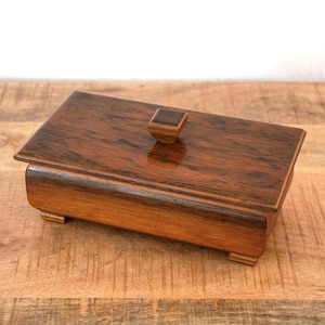 May include: A rectangular wooden box with a dark brown, polished finish and visible wood grain patterns. The box features a flat lid with a small, square-shaped central knob and rests on four short, integrated feet at the base corners.