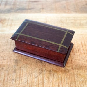 Op de afbeelding: Een rechthoekige houten doos met een donkerbruine afwerking en messing inlegwerk op het deksel. De doos staat op een licht verhoogde basis, met een klassiek ontwerp. De houtnerf is zichtbaar, wat bijdraagt aan de natuurlijke uitstraling.