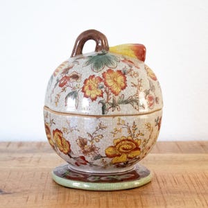 May include: A ceramic lidded jar with a floral design in yellow, orange, and red on a cream backdrop. It features a brown handle and rests on a round base with a green and brown border. The jar is likely for storage, with a height of approximately 15 cm.