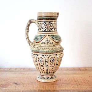 Może przedstawiać: Ozdobny ceramiczny dzban z uchwytem i wylewką. Dzbanek posiada skomplikowane wzory kwiatowe i geometryczne w kolorze beżowym, brązowym, zielonym i niebieskim. Dzbanek stoi na podstawie i jest prezentowany na drewnianej powierzchni.