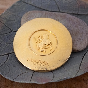 Puede incluir: Un estuche compacto dorado con el logotipo de Lancôme Paris. El estuche compacto presenta un diseño en relieve de un querubín dentro de un marco circular. El estuche compacto es redondo y tiene una superficie texturizada.
