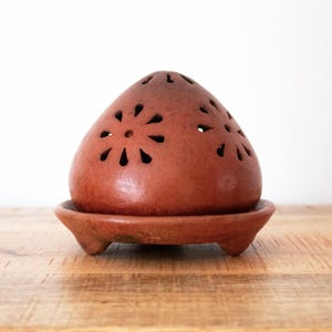 May include: A terracotta incense burner with a domed top and a matching base. The top features decorative cut-out floral patterns. The base has three small feet. The burner is a warm reddish-brown color and sits on a wooden surface.