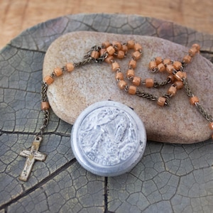 May include: A vintage rosary featuring peach-coloured beads and a silver cross. A small, round, silver container with a relief image and the word "Lourdes" is visible. The items are placed on a stone and a textured, grey surface.