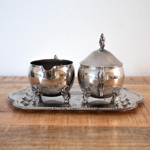 Op de afbeelding: Een zilverkleurige set van een roomstel en suikerpot met een dienblad. De roomstel heeft een tuit en de suikerpot heeft een deksel met een decoratieve knop. Beide staan op sierlijke poten en op een bijpassend dienblad.