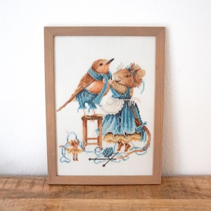 Puede incluir: Obra de arte de punto de cruz en un marco de madera. La imagen muestra un pájaro y un ratón, ambos con ropa azul, participando en una actividad artesanal. El pájaro está posado en un pequeño taburete, y el ratón sostiene hilo. La paleta de colores incluye marrón, azul y blanco.