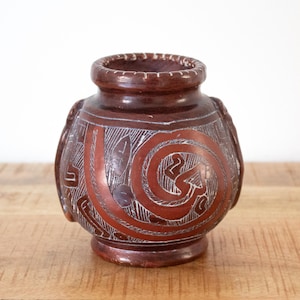 May include: A brown ceramic vase with detailed white etched patterns. The vase has a wide body, a narrow neck, and a swirling design. The vase is displayed on a wooden surface.